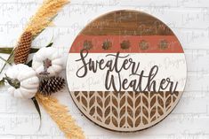 a wooden sign with the words sweater weather on it next to some flowers and pine cones