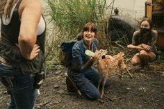 two women and one man wearing masks are playing with a baby deer in the dirt