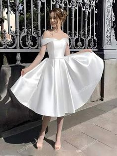 a woman in a white dress standing on the sidewalk with her hands behind her back