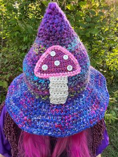 a woman with pink hair wearing a crocheted hat and purple shirt in front of bushes