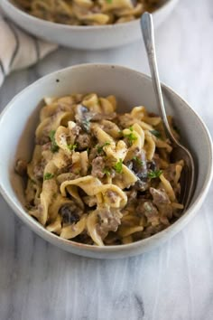 two bowls filled with pasta and meat on top of a marble countertop next to a silver spoon