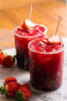 two glasses filled with liquid and strawberries on top of a marble table next to each other