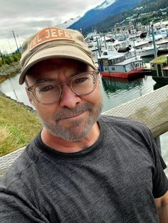 a man wearing glasses and a hat standing in front of a harbor filled with boats