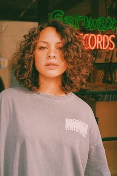 a woman with curly hair standing in front of a store window wearing a gray shirt