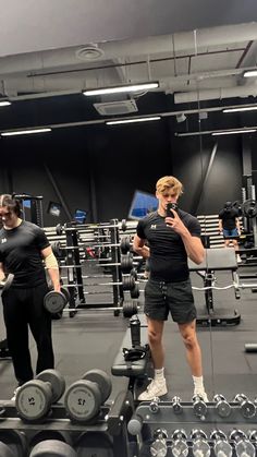 two men are in the gym with dumbbells and one is looking at his cell phone