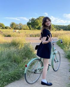 a woman standing next to a bike on a dirt road with grass and trees in the background