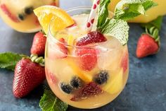 two glasses filled with fruit and ice on top of a blue table next to lemons, strawberries, and mint