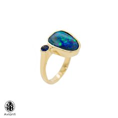 "Surprise her with this stunning blue boulder opal ring. The center stone is an squarish Pear cut natural boulder Australian Opal and is cast in 14 Karat yellow gold, with shiny finish with an accent of bezel set Sapphire stone. This ring is from our \"Opalescence \" collection. Each stone is unique in cut and color. This beautiful stone displays a green and blue hue that compliment each other in every way. [MATERIALS] Stone: Opal (1) Cut: Pear Carat: 3.03 Play of color: blue green hues Transpar Blue Cabochon Teardrop Ring, Blue Teardrop Cabochon Rings, Formal Blue Opal Rings, Blue Cabochon Opal Ring For Anniversary, Anniversary Blue Cabochon Opal Ring, Blue Opal Cabochon Ring, Blue Opal Ring With Bezel Setting For Gift, Unique Opal Ring, Yellow Gold Opal Ring