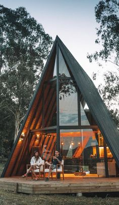 two people sitting on chairs in front of a wooden cabin with glass walls and windows