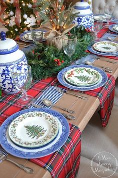 the table is set for christmas dinner with blue and white plates, silverware, greenery, and a potted tree