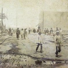 an old black and white photo of people walking on a dirt road with buildings in the background