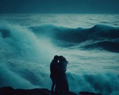 two people standing on rocks near the ocean with their arms around each other as waves crash in the background