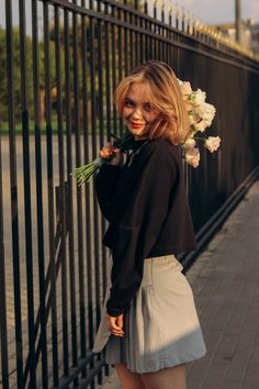 a woman standing next to a fence with flowers in her hair and wearing a skirt