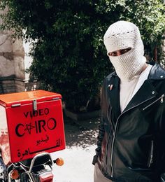 a man wearing a white mask standing next to a motorcycle with a red box on it