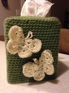 a green crocheted tissue holder with two white flowers on it, sitting on a table