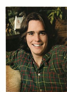 a young man with long hair wearing a green and red plaid shirt smiles at the camera