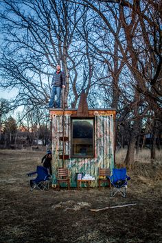 a man standing on top of a small building in the middle of a field next to two lawn chairs