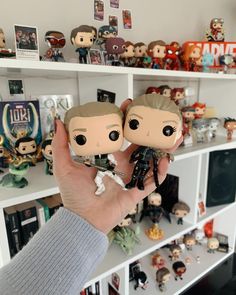 two figurines are held up in front of a shelf full of pop figures