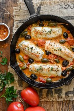 fish with olives and tomatoes in a skillet on a wooden table next to fresh herbs