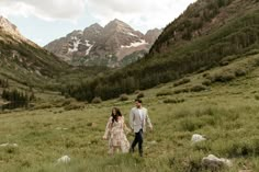 a man and woman walking through a lush green field with mountains in the back ground