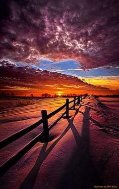 the sun is setting over a snowy field with a fence and trees in the foreground