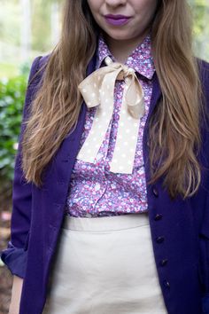 a woman with long hair wearing a purple jacket and white skirt, standing in front of some bushes