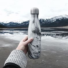 a person holding up a water bottle in front of a body of water with mountains in the background