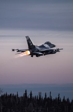 a fighter jet flying through the air with a light on it's side and trees in the background