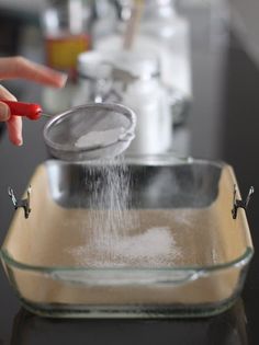 a person is sprinkling flour in a glass dish on a counter top with a red spatula