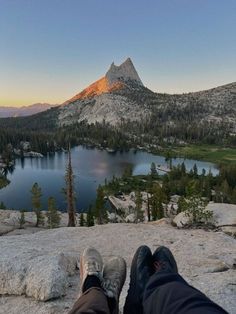 someone is sitting on the edge of a cliff overlooking a lake and mountain range at sunset