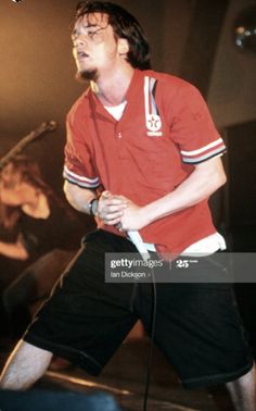 a man in red shirt and black shorts holding a microphone on stage with other people behind him