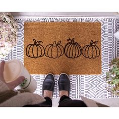 a person standing in front of a door mat with three pumpkins on it and a coffee cup