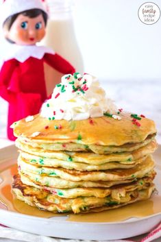 an elf is standing next to a stack of pancakes with whipped cream and sprinkles