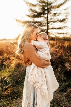 a woman holding a baby in her arms while walking through the grass with trees in the background