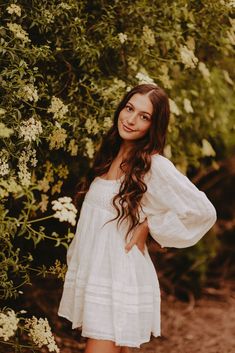 a beautiful young woman standing in front of some trees and flowers wearing a white dress