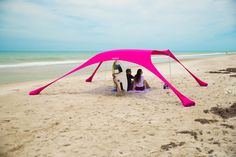 three people are sitting under a pink tent on the sand at the beach near the ocean