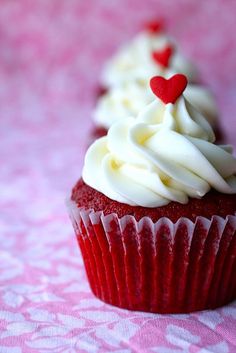 two red velvet cupcakes with white frosting and hearts on top, sitting on a pink tablecloth