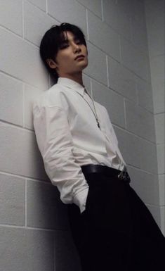 a man leaning against a wall wearing a white shirt and black pants with his hands on his hips