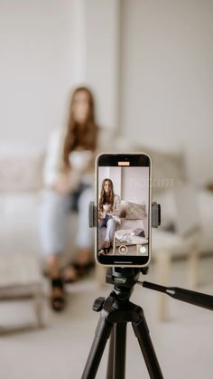 a woman sitting on a couch behind a tripod taking a photo with her cell phone