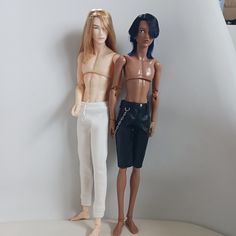 two dolls standing next to each other on top of a white surface with a wall in the background