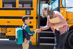 an older woman is shaking hands with a young boy in front of a school bus