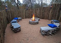 an outdoor fire pit with benches and pillows on the ground in front of a bamboo fence