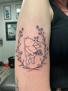 A line work tattoo of a traditional Winnie the pooh as drawn by E. H. Shepard holding a bundle of violets and surrounded by a wreath of peonies positioned on the left bicep. American Traditional Winnie The Pooh Tattoo, Classic Pooh And Piglet, Vintage Pooh Tattoo, Winnie And Eeyore Tattoo, Tigger And Pooh Tattoo, Subtle Winnie The Pooh Tattoo, Vintage Winnie The Pooh Tattoo, Winnie The Pooh Outline Tattoo, Classic Pooh Tattoo