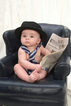 a baby sitting in a chair with a hat on and reading a newspaper while wearing a tie