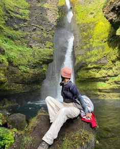 a man sitting on top of a rock next to a waterfall in the forest with a red backpack