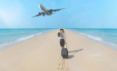 a woman is standing on the beach with her suitcase looking at an airplane flying overhead