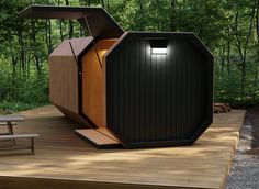 an outdoor toilet on a wooden deck surrounded by trees and grass, with a bench in the foreground