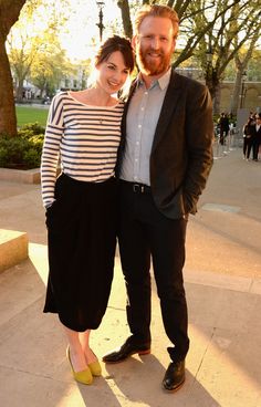 a man and woman standing next to each other in front of some trees on a sunny day