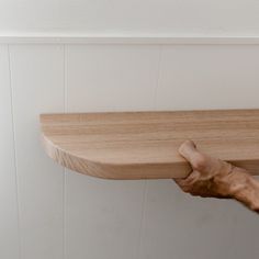 a person holding onto a wooden shelf with their thumb on the edge, in front of a white wall