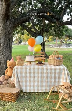 a picnic table with balloons and teddy bears in the grass under a large oak tree